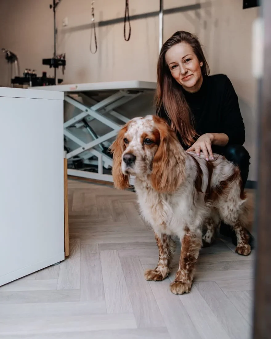 Alicja, założycielka Ginger Dog, kucająca w studiu przy swoim cocker spanielu Winstonie, za nimi stół groomerski